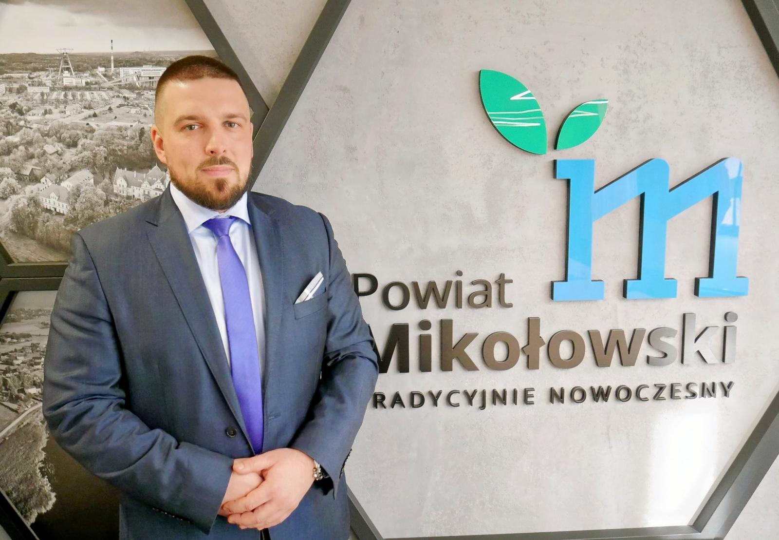 Prezes Centrum Zdrowia w Mikołowie Mateusz Niewdana stoi na tle logo Powiatu Mikołowskiego